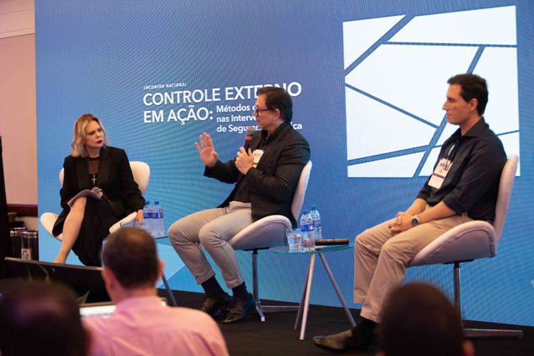 Três pessoas estão sentadas em cadeiras brancas em um palco durante um evento. No centro, um homem de óculos e paletó preto fala ao microfone. À esquerda, uma mulher loira de preto ouve segurando outro microfone. À direita, um homem de camisa azul ouve com as mãos no colo. Atrás deles, há uma grande tela azul com o texto "CONTROLE EXTERNO EM AÇÃO" e um logotipo branco. Algumas pessoas da plateia aparecem no primeiro plano assistindo à apresentação.