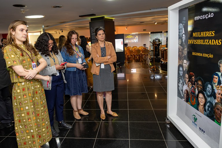 A fotografia mostra quatro mulheres em pé, lado a lado, observando atentamente um grande painel expositivo à direita em um ambiente interno de piso escuro e brilhante.
