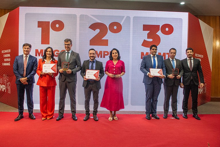 A imagem mostra uma cerimônia de premiação em um palco. No centro, há um painel digital grande com três posições numeradas, "1º," "2º," e "3º," indicando as classificações. Da esquerda para a direita, há três grupos de pessoas. Todos estão vestidos formalmente, e a atmosfera é de celebração.