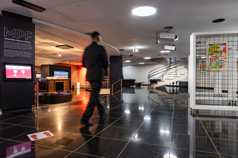 Fotografia em plano geral do Espaço Memorial do Ministério Público Federal (MPF), um ambiente interno amplo, moderno e bem iluminado.   No centro da imagem, um homem vestindo um fato escuro caminha de costas para a esquerda, ligeiramente desfocado devido ao movimento. O chão é revestido por ladrilhos de granito preto.

À esquerda, destaca-se um painel preto de exposição com a sigla "MPF" em letras grandes no topo, seguida do texto explicativo "MINISTÉRIO PÚBLICO FEDERAL". Abaixo, há um ecrã digital integrado. 

Ao fundo, à direita, encontra-se uma escada em caracol de metal escuro com degraus claros e corrimão, que dá acesso ao piso superior. No teto branco, observam-se várias luminárias embutidas e um projetor suspenso.   

À direita, em primeiro plano, vê-se uma estrutura branca com uma grelha metálica onde está exposta uma obra de arte colorida em estilo expressionista/urbano.