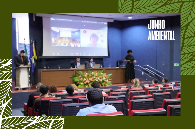 Foto com pessoas em um auditório assistindo a um evento com palestrantes, telão e intérprete de Libras no palco. Imagem está inserida numa moldura verde com o texto Junho Ambiental no canto direito superior