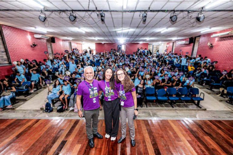 Foto tirada durante uma apresentação do projeto Manda a Real em uma escola. Em primeiro plano, no centro da imagem, estão três integrantes do projeto, duas mulheres e um homem, sorrindo para a câmera. Eles usam as camisetas roxas do Manda a Real e estão com crachás de identificação. Ao fundo, há uma grande plateia de estudantes reunida no espaço da escola. Muitos estão sentados em cadeiras, enquanto outros permanecem em pé; eles usam uniformes azuis. Vários estudantes olham para a câmera, acenam ou fazem gestos de comemoração. O ambiente tem paredes vermelhas e um piso de madeira, e a cena transmite um clima de entusiasmo e participação durante o evento.