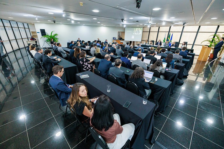 Pessoas em traje formal participam num evento num auditório espaçoso, dispostas em mesas cobertas com toalhas pretas viradas para uma mesa principal com bandeiras e um ecrã de projeção ao fundo.