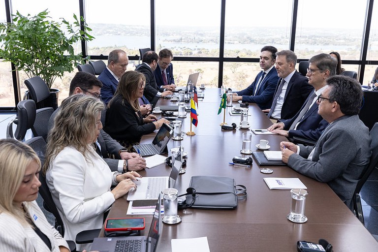Foto mostra homens e mulheres vestidos formalmente sentados em torno de mesa comprida em sala de reunião. Alguns deles têm notebooks à frente, sobre a mesa, e há também pequenas bandeiras de países sobre a mesa