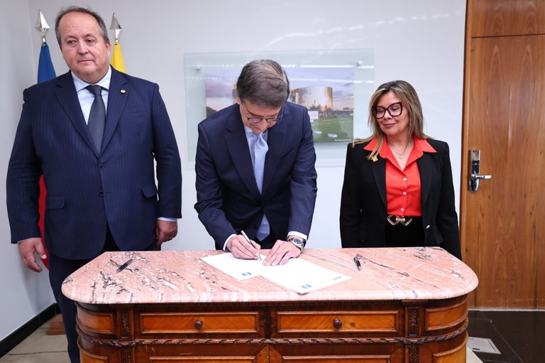 Homem de terno assina um documento sobre mesa de mármore rosa. Ao seu lado, outro homem de terno e uma mulher de terno preto e camisa vermelha observam o ato formal.
