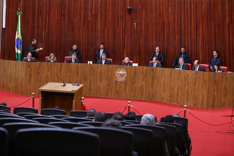Foto do plenário do Tribunal Superior Eleitoral, com ministros ou juízes sentados em uma bancada elevada com o emblema da República, em um plenário com painéis de madeira e uma plateia em primeiro plano.