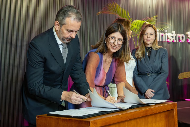 Fotografia colorida de Raquel Branquinho e Alexandre Espinosa inclinados sobre um púlpito de madeira enquanto assinam documentos com canetas.