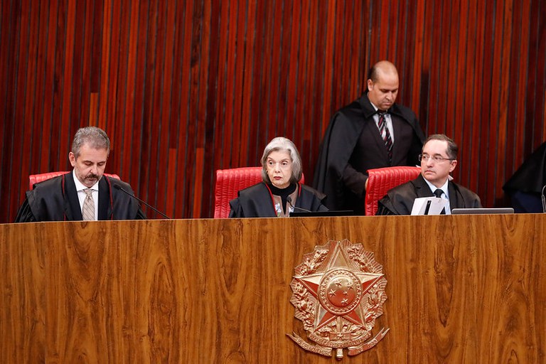 Fotografia de ministros do TSE em suas cadeiras no plenário do tribunal, com a ministra Cármen Lúcia ao centro, com a parede do local e a bancada revestidos de madeira e o brasão da República no centro da bancada.