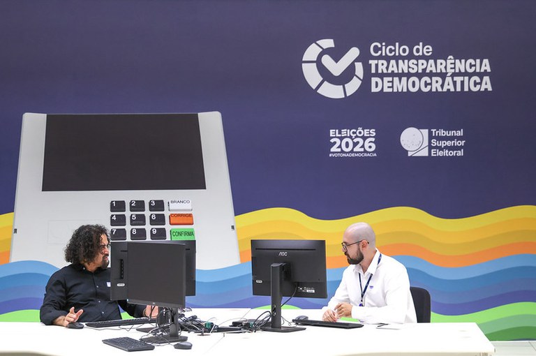 A foto mostra dois homens sentados em frente a computadores. Ao fundo da foto, há um painel escrito "Ciclo de Transparência Democrática, Eleições 2026 e Tribunal Superior Eleitoral".
