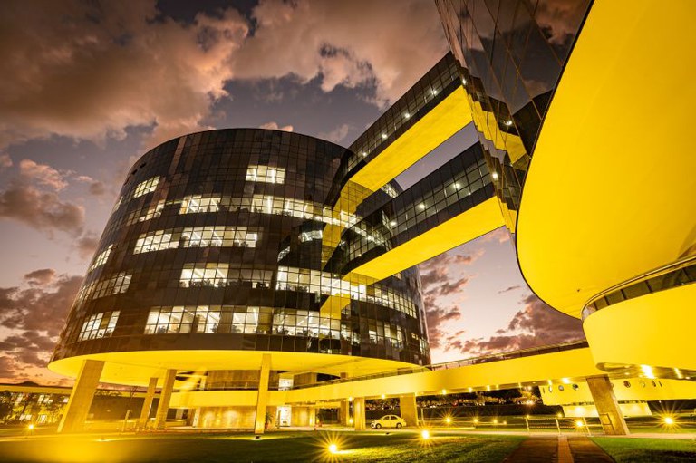 Foto dos prédios da PGR ao entardecer. Os prédios estão iluminados em amarelo. É possível ver algumas salas com as luzes acessas.