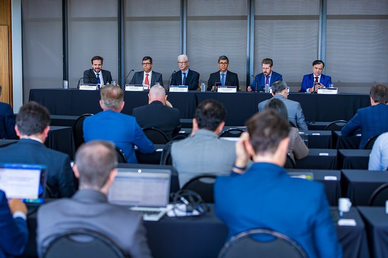 Mesa com seis homens de terno e gravata em um painel de discussão, de frente para uma plateia vista de costas em um auditório.