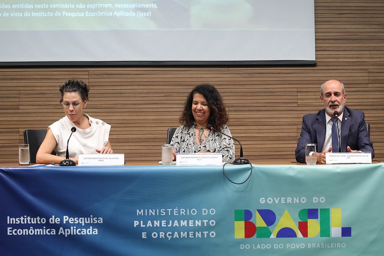 Foto mostra duas mulheres e um homem atrás de uma bancada, fazendo uma apresentação.