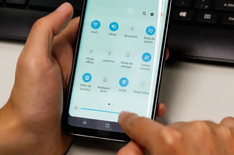 Close-up de mãos segurando um smartphone que exibe um menu de configurações rápidas com diversos ícones, como Wi-Fi e Bluetooth, em frente a um teclado de computador desfocado.