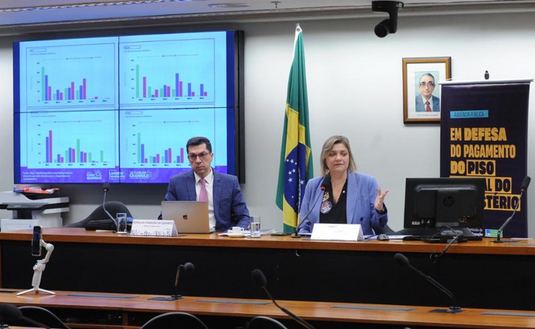 Fotografia colorida de uma audiência pública. À esquerda, um homem com terno e óculos olha para frente. À direita, uma mulher com blazer azul e cabelo loiro gesticula enquanto fala. Ao fundo, um telão exibe gráficos de barras, e uma faixa preta exibe as palavras "AUDIÊNCIA PÚBLICA EM DEFESA DO PAGAMENTO DO PISO SALARIAL DO MAGISTÉRIO".
