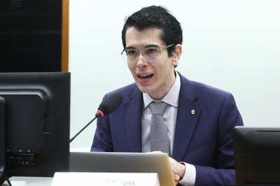 O procurador da República André Luiz Porreca representa o MPF na audiência pública "Impactos do uso do mercúrio em trabalhadores", realizada pela Câmara dos Deputados, em 05/05/2026. Foto: Vinicius Loures/Câmara dos Deputados. A imagem apresenta um plano médio de um homem jovem, de pele clara e cabelos escuros penteados para trás, em um ambiente formal que sugere uma audiência ou conferência. Ele utiliza óculos de grau com armação fina, veste um paletó azul-marinho sobre uma camisa social clara e uma gravata com padronagem geométrica cinza, ostentando um pequeno broche dourado na lapela. O homem está com a boca levemente aberta, capturado no momento em que fala ao microfone posicionado à sua frente. Ao redor dele, é possível ver parte de um monitor de computador à esquerda e o topo de uma placa de identificação na base da foto, reforçando o cenário institucional de debate ou pronunciamento oficial.