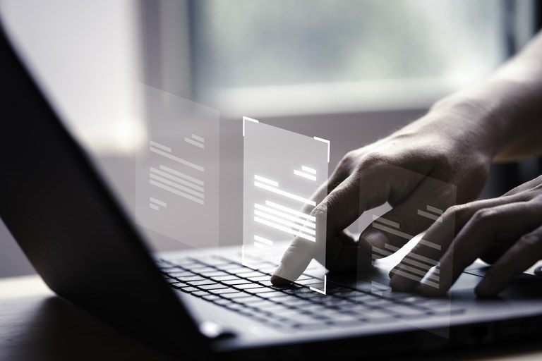 Mão usando laptop com tela virtual. Ao fundo, há uma janela.