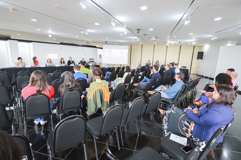 sala ampla com cadeiras pretas, pessoas sentadas nas cadeiras e uma mesa grande à frente, com microfones e pessoas falando ao microfone
