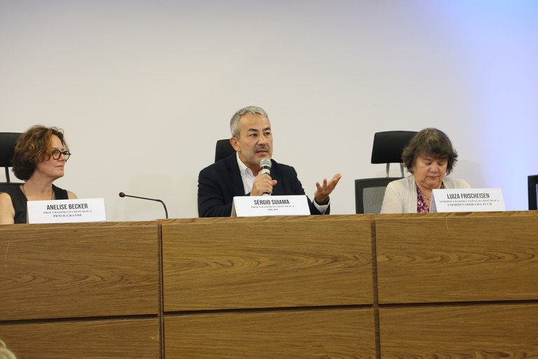 imagem de mesa larga com um homem ao centro e duas mulheres à esquerda e à direita. o homem segura o microfone.