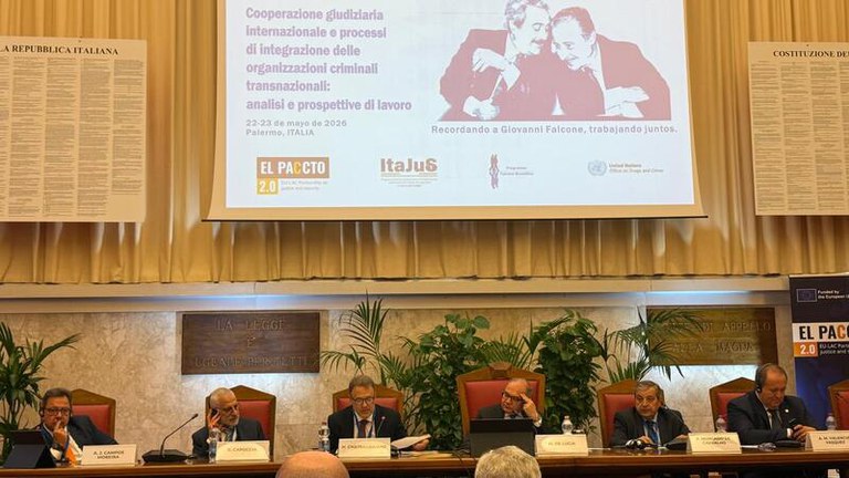 A imagem mostra um painel com seis homens de terno sentados em uma longa bancada durante um evento formal. Atrás deles, há um grande telão que exibe o título de uma conferência sobre cooperação judiciária internacional e crime organizado, realizada em Palermo em maio de 2026. O telão também destaca logos institucionais e uma ilustração dos juízes italianos Giovanni Falcone e Paolo Borsellino.