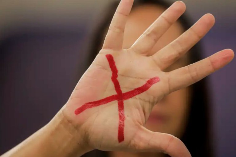 Foto de uma mão com um X vermelho em primeiro plano. Ao fundo, desfocado, é possível ver uma mulher de pele branca e cabelos escuros.