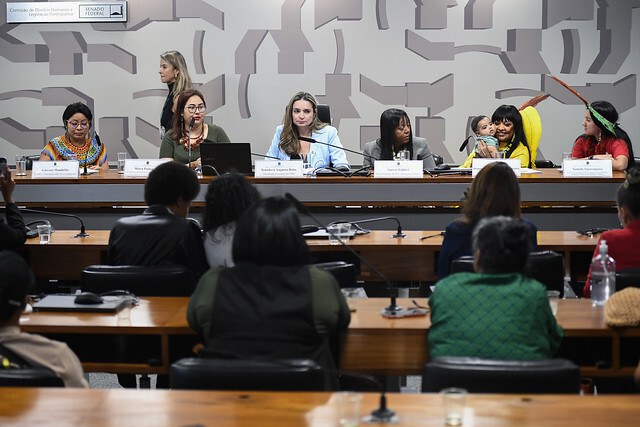Imagem mostra seis mulheres, sendo 5 delas indígenas, sentadas atrás de uma bancada. Em frente à elas, está o público que assiste à audiência.