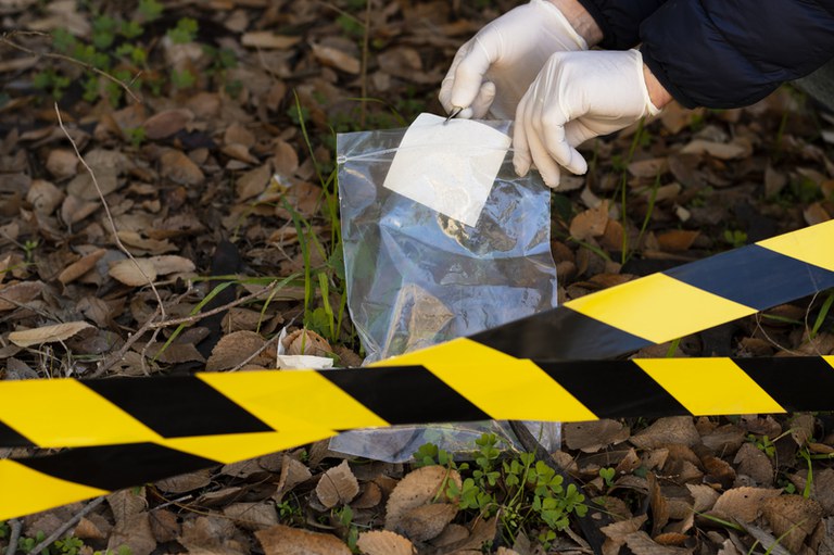 Uma pessoa usando luvas brancas segura um saco plástico transparente de recolhimento de evidências em uma área externa cercada por uma fita de isolamento amarela e preta. Ao fundo, o chão está coberto de folhas secas e pouca vegetação.