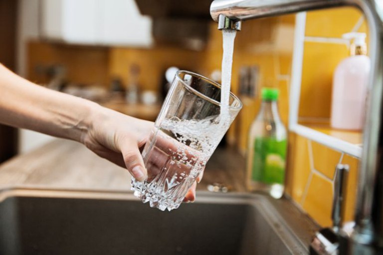 Foto mostra uma pessoa segurando um copo e enchendo de água na torneira de uma pia de cozinha