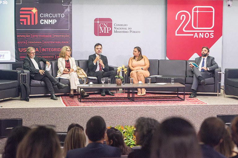 A imagem mostra o procurador da República Daniel Azeredo sentado à mesa de um painel, com microfone à frente, gesticulando enquanto fala. À mesa, há outros especialistas e autoridades, alguns com laptops e documentos, atentos à discussão. Ao fundo, é possível ver uma tela de projeção ou banner com o tema do painel, “O Brasil na trilha da descarbonização: atuação estratégica para o equilíbrio climático”. O ambiente é um auditório moderno, com iluminação clara, cadeiras organizadas para o público e uma mesa central longa com copos de água, microfones e materiais de apoio.