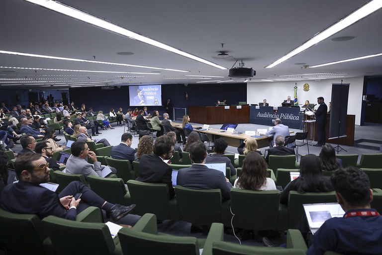 Foto do plenário da 2ª Turma do STF que recebeu a audiência pública sobre emendas impositivas. Mostra o plenário, de cadeiras verdes, cheio com plateia. É possível também ver a mesa com as pessoas que falaram na audiência pública e as autoridades. É possível ler o banner de mesa "audiência pública"