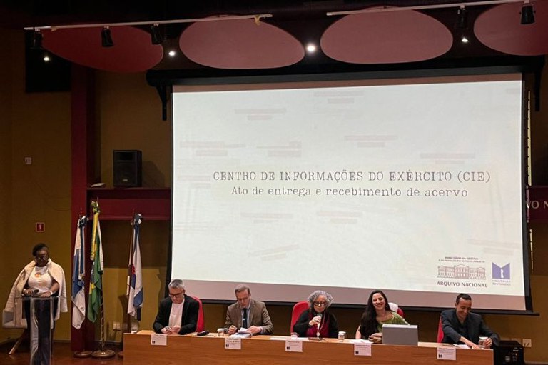 Foto mostra mesa do evento com cinco pessoas sentadas e uma em pé no púlpito ao lado; por trás da mesa, em um telão, está escrito Centro de Informações do Exército CIE ato de entrega e recebimento de acervo