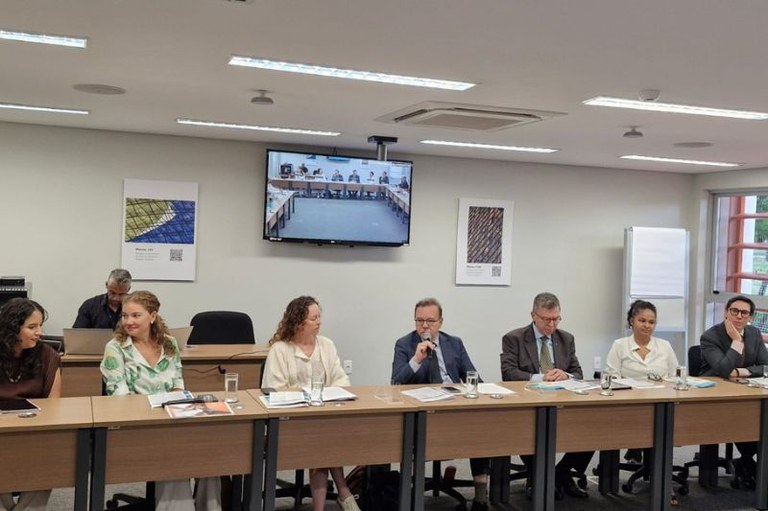 Foto mostra uma mesa com oito pessoas e um telão atrás