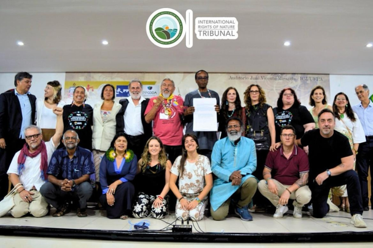 A imagem mostra um grande grupo de pessoas posando para uma foto em um palco. O grupo é diverso, composto por cerca de vinte pessoas de diferentes etnias e idades, com alguns em pé na parte de trás e outros agachados na frente. Acima do grupo, há um logotipo e texto em inglês que diz "INTERNATIONAL RIGHTS OF NATURE TRIBUNAL" (Tribunal Internacional dos Direitos da Natureza), indicando o contexto do evento.