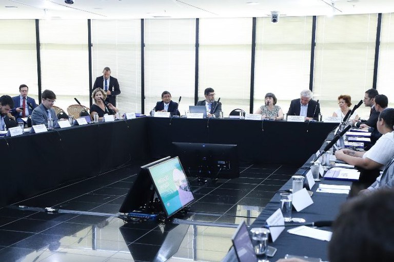 A imagem mostra uma reunião formal em uma sala bem iluminada, com várias pessoas sentadas ao redor de uma mesa preta em formato de "U". Os participantes estão vestidos de maneira formal, com ternos, blazers e roupas sociais. Alguns estão usando óculos e há uma diversidade de idades entre os presentes. Uma mulher de cabelos curtos, vestindo uma blusa estampada, segura um microfone e parece estar falando. Outras pessoas ao redor tomam notas, digitam em laptops ou seguram documentos.