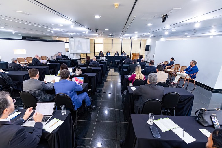 A imagem mostra um evento profissional de negócios, possivelmente um workshop ou reunião, com participantes assistindo à uma apresentação em uma sala de conferências.
