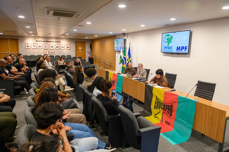 A imagem mostra um auditório durante um evento do Ministério Público Federal (MPF), com foco na mesa diretora onde quatro palestrantes se reúnem em frente às bandeiras do Brasil e do DF, e uma tela com os logos "PFDC" e "MPF". À frente da mesa, uma grande faixa tricolor com o nome "EDUCAFRO" está estendida, simbolizando a temática do encontro. A plateia, majoritariamente composta por pessoas negras, ocupa as poltronas.o.
