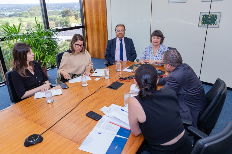 A imagem mostra seis pessoas reunidas em uma sala de conferência ao redor de uma mesa de madeira grande. Elas estão participando de uma reunião e parecem concentradas. A sala tem grandes janelas que deixam entrar bastante luz natural e mostram uma vista com árvores e um campo verde ao fundo