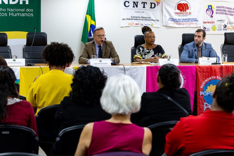 Foto mostra auditório do evento com bandeiras das instituições penduradas na parede ao fundo, no canto superior esquerdo o banner do CNDH - conselho nacional dos direitos humanos, à frente, uma mesa com dois homens de terno e uma mulher negra ao centro, outras bandeiras penduradas na parte da frente da mesa e de frente para a mesa o público que acompanhou