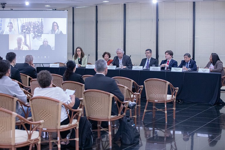 Foto de auditório com mesa de debatedores ao fundo e um telão à esquerda, com pessoas na tela