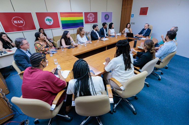Imagem mostra uma reunião presencial ao redor de uma grande mesa de madeira retangular. Cerca de 15 pessoas estão sentadas em cadeiras de escritório, acompanhando a fala de um homem de terno posicionado na cabeceira da mesa. Na parede ao fundo, estão pendurados quadros com bandeiras e logotipos de movimentos sociais
