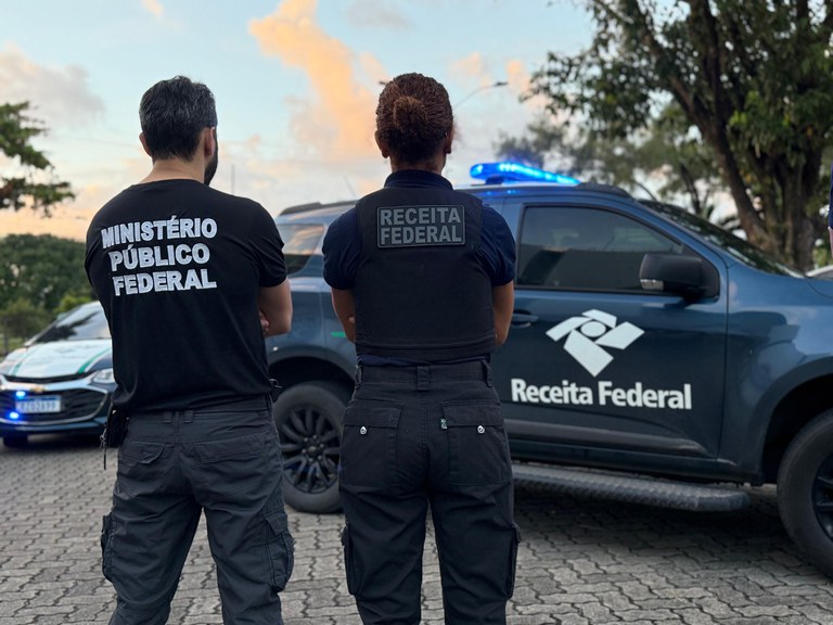 Dois agentes de costas, do Ministério Público Federal e da Receita Federal, observam uma viatura policial caracterizada da Receita Federal com giroflex ligado.