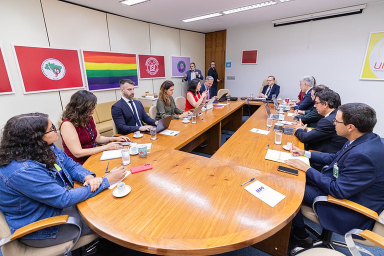 A imagem mostra uma reunião em torno de uma grande mesa de madeira em formato de "U", com cerca de doze participantes sentados em cadeiras de escritório. A maioria das pessoas veste trajes formais, manuseia laptops e papéis, enquanto o ambiente exibe quadros coloridos na parede de fundo.