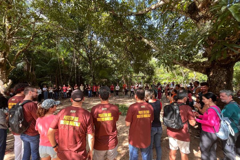 Foto tirada ao ar livre, em uma área com muitas árvores e chão de terra batida, onde um grupo de cerca de 25 pessoas está reunido. A maioria observa atentamente uma mulher que fala em primeiro plano, vestindo camiseta marrom com os dizeres “Povos e Comunidades Tradicionais VIVEM!”, calça preta e tênis branco. Ela segura um boné branco na mão. À direita, um homem também com a mesma camiseta a observa com as mãos na cintura. Outras pessoas do grupo, de diferentes idades, etnias e estilos de roupa, estão sorrindo, prestando atenção ou conversando entre si.
