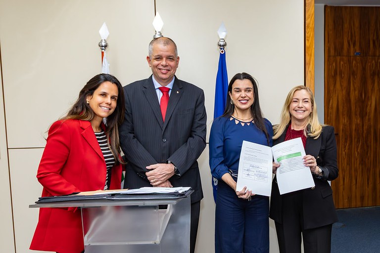 Fotografia de quatro pessoas (Ana Carolina, Robalinho, Giselle Ferreira e Eliana Torelly) em pé posando para a câmera na cerimônia realizada no gabinete da Secretaria-Geral (PGR). Eliana e Giselle seguram o documento assinado.