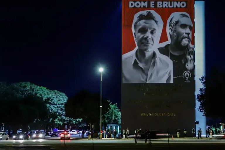 Foto ilustrativa de junho de 2024 - A foto aparece projetada na lateral do ministério dos Direitos Humanos na Esplanada dos Ministérios, em Brasília. A foto está com fundo vermelho e relembra , à época, os dois anos da morte do indigenista Bruno Pereira e do jornalista Dom Phillips. A foto dos dois está em preto e branco.