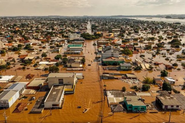 Fotografia aérea de uma cidade amplamente alagada por uma enchente. As ruas estão completamente cobertas por água barrenta, e a inundação atinge casas, comércios e outras construções urbanas. Apenas os telhados e partes superiores dos prédios estão visíveis. Pequenas embarcações podem ser vistas navegando pelas ruas inundadas, indicando que o nível da água é bastante elevado. Ao fundo, a imagem mostra a imensidão da cidade afetada e o horizonte encoberto por nuvens claras.