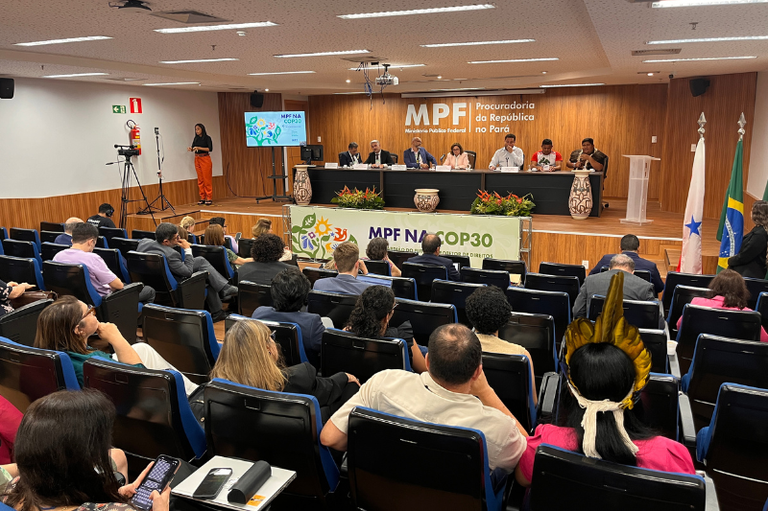 Uma vista de um auditório lotado onde está ocorrendo um evento do MPF na COP30 (Ministério Público Federal). No palco elevado, um painel de sete autoridades está sentado em uma longa mesa, com um grande logo do MPF ao fundo. Uma pessoa, possivelmente indígena, na plateia, usa um cocar amarelo em primeiro plano.