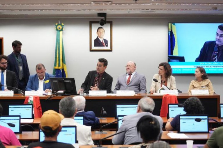 Foto mostra a sala da audiência com o público, a mesa dos palestrantes e o telão
