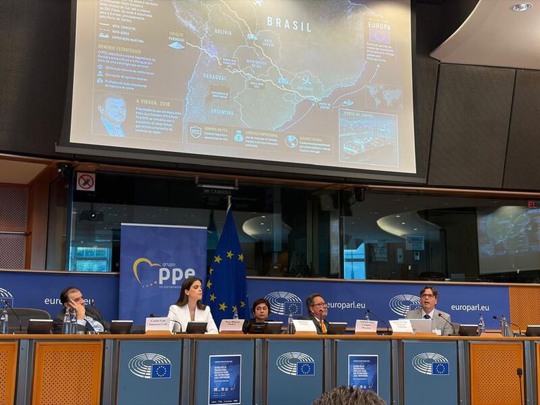 A imagem mostra um painel de discussão no Parlamento Europeu com cinco palestrantes sentados atrás de uma bancada com a bandeira da União Europeia e um banner do Grupo PPE. Acima deles, uma grande tela projeta um mapa e textos em português detalhando as rotas logísticas e o domínio estratégico do crime organizado entre o Brasil, Paraguai e a Europa.