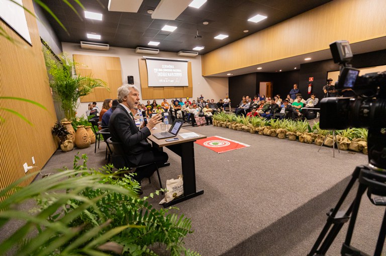 Foto de uma sala moderna e bem iluminada. Um palestrante está sentado à frente com microfone e laptop, falando para uma plateia atenta. Há plantas decorativas, vasos grandes e uma câmera gravando o evento.