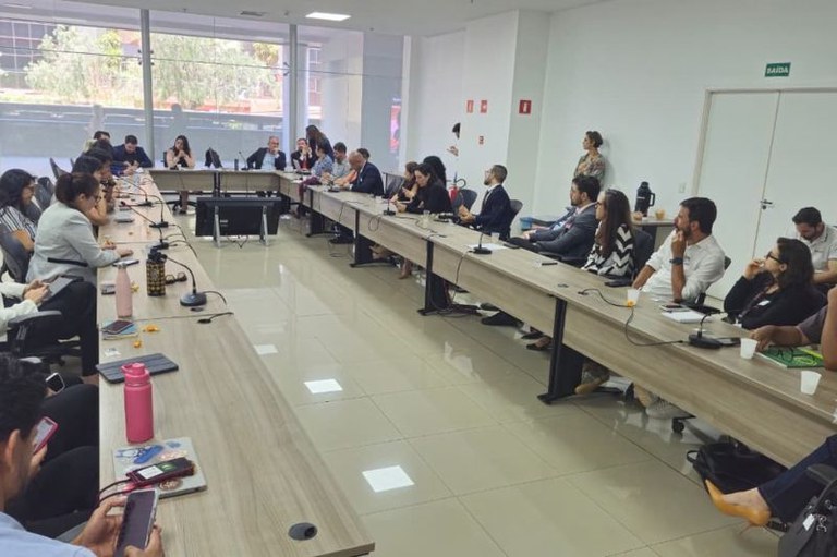 Foto mostra uma sala de reuniões com uma mesa em U com microfones, cadernos, celulares garrafas de água e diversas pessoas entre homens e mulheres sentadas; a ala tem uma janela grande ao fundo e ao lado uma porta está fechada